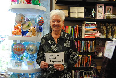 This is a picture of a older female smiling and holding a sign that says:
"I am the patient Experience!"