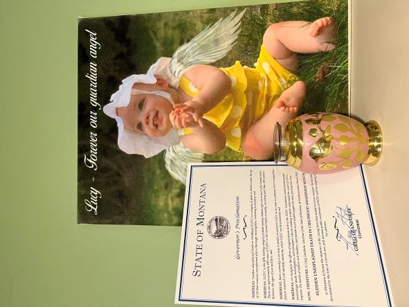 Picture of a paper that says : State of Montana 
Governor&apos;s Proclamation &quot;SUDDEN UNEXPLAINED DEATH IN CHILDHOOD AWARENESS MONTH&quot; 
Picture of baby Lucy sitting outside in the grass wearing a hat. She has her hands together, clapping and smiling while looking up. There is angel wings on her backside.
Picture of a pretty small urn of her ashes that has a little bird on it.
March is SUDC Awareness Month, and Montana is one of the many states that has recognized it as such in recent years.