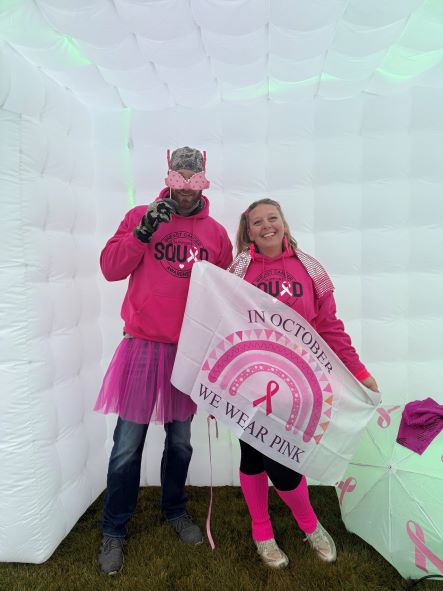 Man and Woman dressed in all pink. The female is holding a flag that says In October We Wear Pink. It has a rainbow with a breast cancer ribbon.